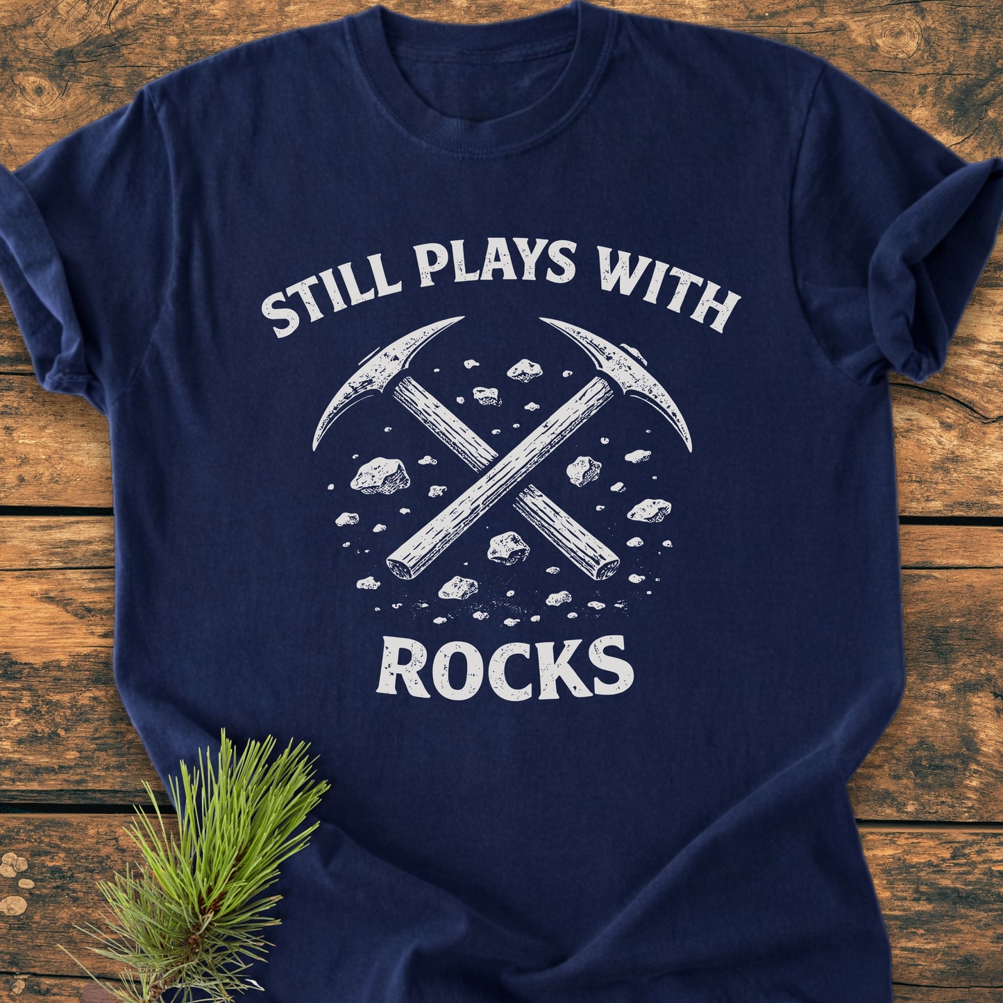 Navy-blue Still Plays With Rocks - Vintage Wash Tee featuring white print and crossed pick-axes design, laid flat on rustic wood with a pine branch accent, highlighting its relaxed fit and durability.