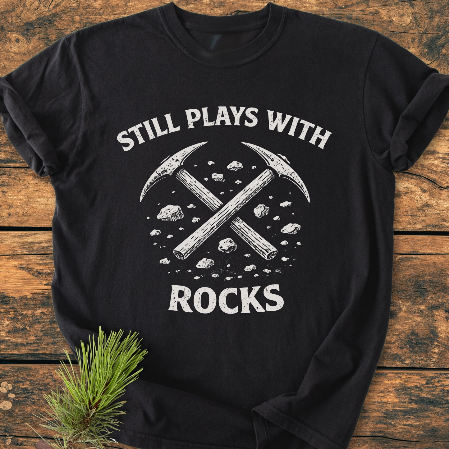 Still Plays With Rocks - Vintage Wash Tee features crossed pick-axes with rocks on a black T-shirt, laid flat on wood. A pine sprig complements the design, emphasizing durability and comfort.