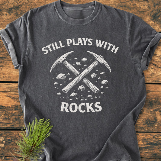 Dark-gray Still Plays With Rocks - Vintage Wash Tee featuring crossed pick-axes and stones, laid on wood. Styled with a pine sprig, emphasizing its relaxed fit and durable, soft-washed fabric.