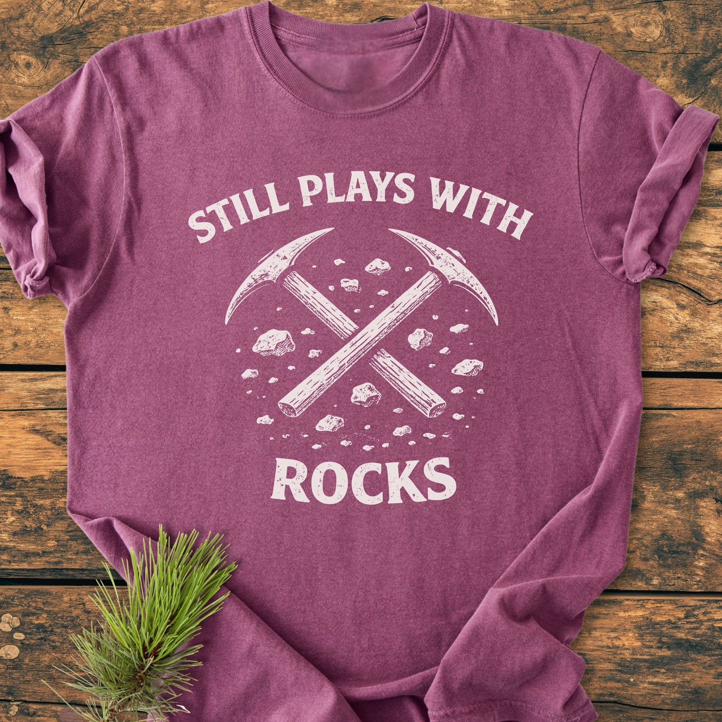 Folded Still Plays With Rocks - Vintage Wash Tee featuring white crossed pickaxes graphic and slogan, laid on wood. A pine sprig accents the scene, highlighting the tee's comfort and design appeal.