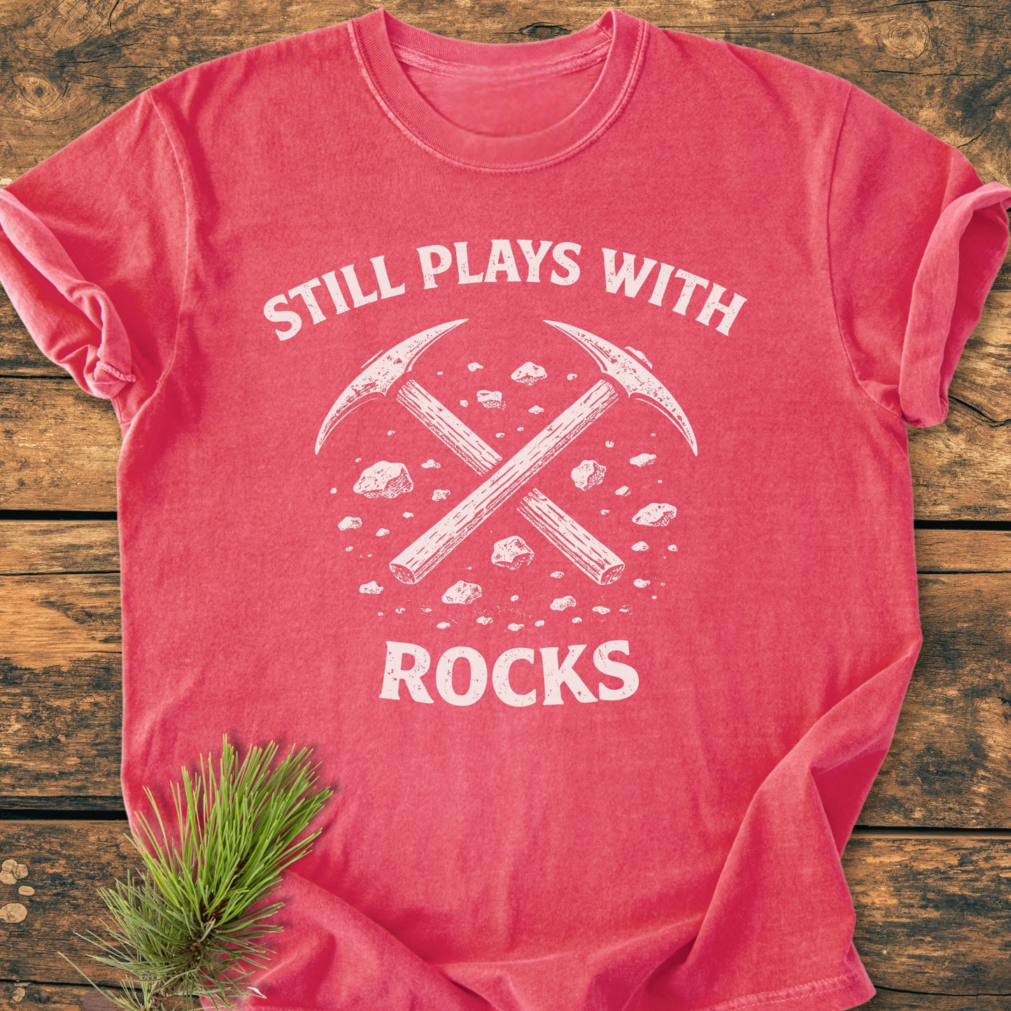 Still Plays With Rocks - Vintage Wash Tee, featuring a white crossed pick-axes graphic and slogan on a red shirt, laid on wood with a pine sprig beside it.