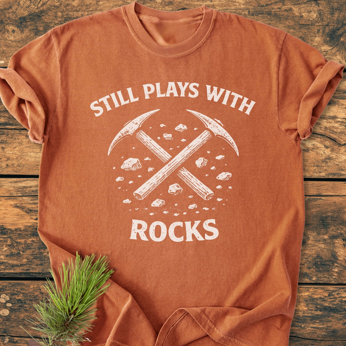 Still Plays With Rocks - Vintage Wash Tee features a white graphic of crossed rock-picks on an orange-rust T-shirt, laid flat with a pine sprig beside it, highlighting its relaxed fit and durability.