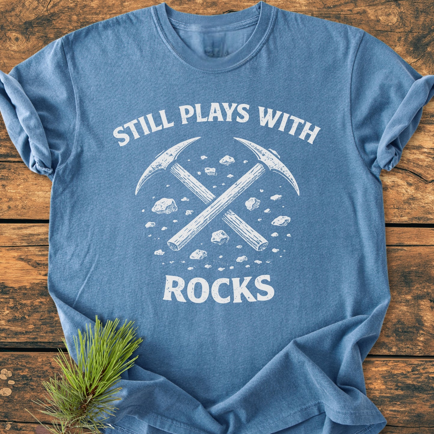 Still Plays With Rocks - Vintage Wash Tee features a heather-blue color with crossed pick-axes and rock fragments printed in white, laid flat with a pine sprig beside it.