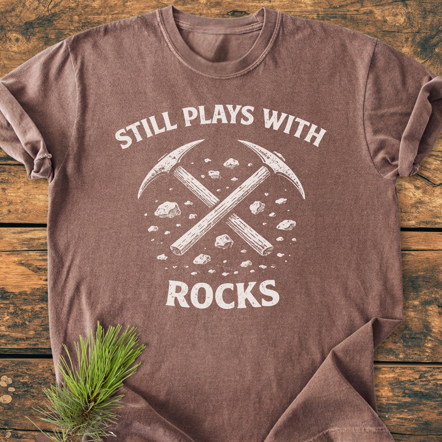 Still Plays With Rocks - Vintage Wash Tee features white crossed pickaxes and stones print on a brown T-shirt, laid on wood with a pine sprig, highlighting its rugged charm and relaxed fit.