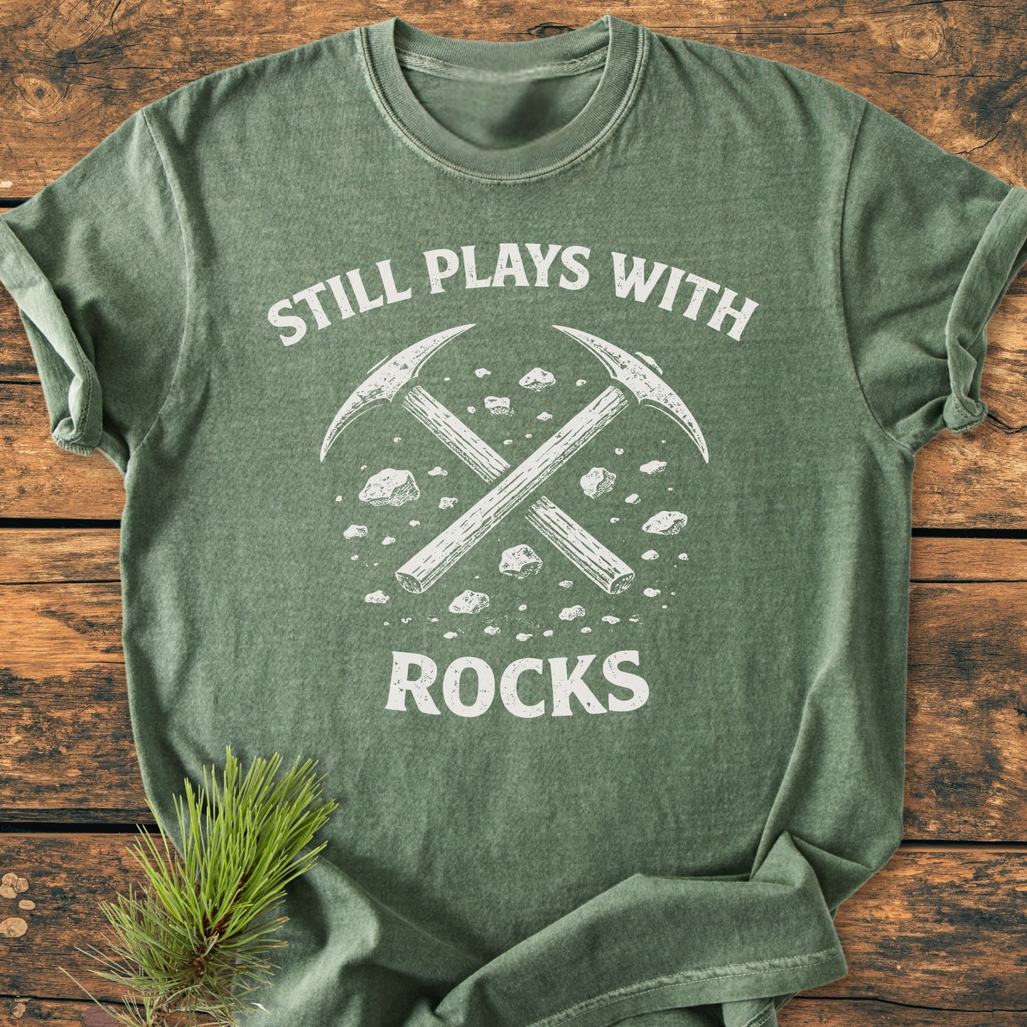 Still Plays With Rocks - Vintage Wash Tee in heathered forest-green, featuring white text and rock-picks graphic. Laid flat on wood, highlighting soft-washed fabric, relaxed fit, and durable stitching.