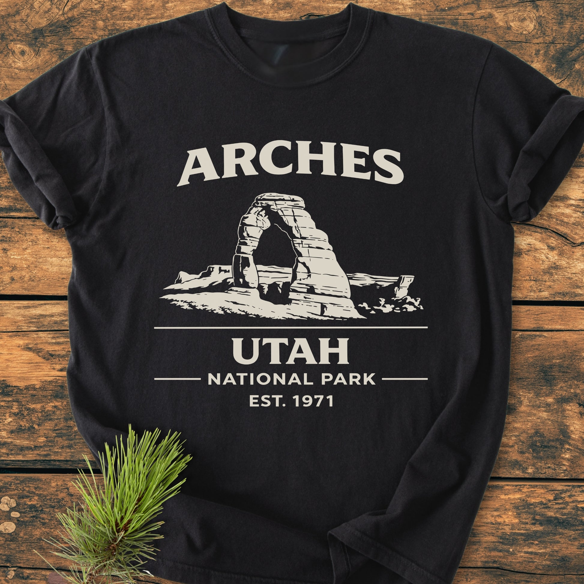 Arches National Park - Vintage Wash Tee featuring Delicate Arch outline and text, laid flat on wood with a pine sprig; made of soft-washed, pre-shrunk 100% ring-spun cotton for comfort.