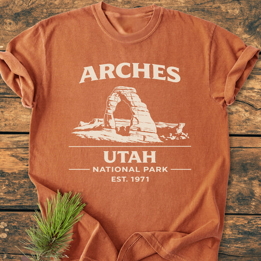 Arches National Park - Vintage Wash Tee featuring a white, vintage-style graphic on a burnt-orange T-shirt, showcasing Delicate Arch with text ARCHES, UTAH NATIONAL PARK, EST. 1971, laid on a wooden surface.