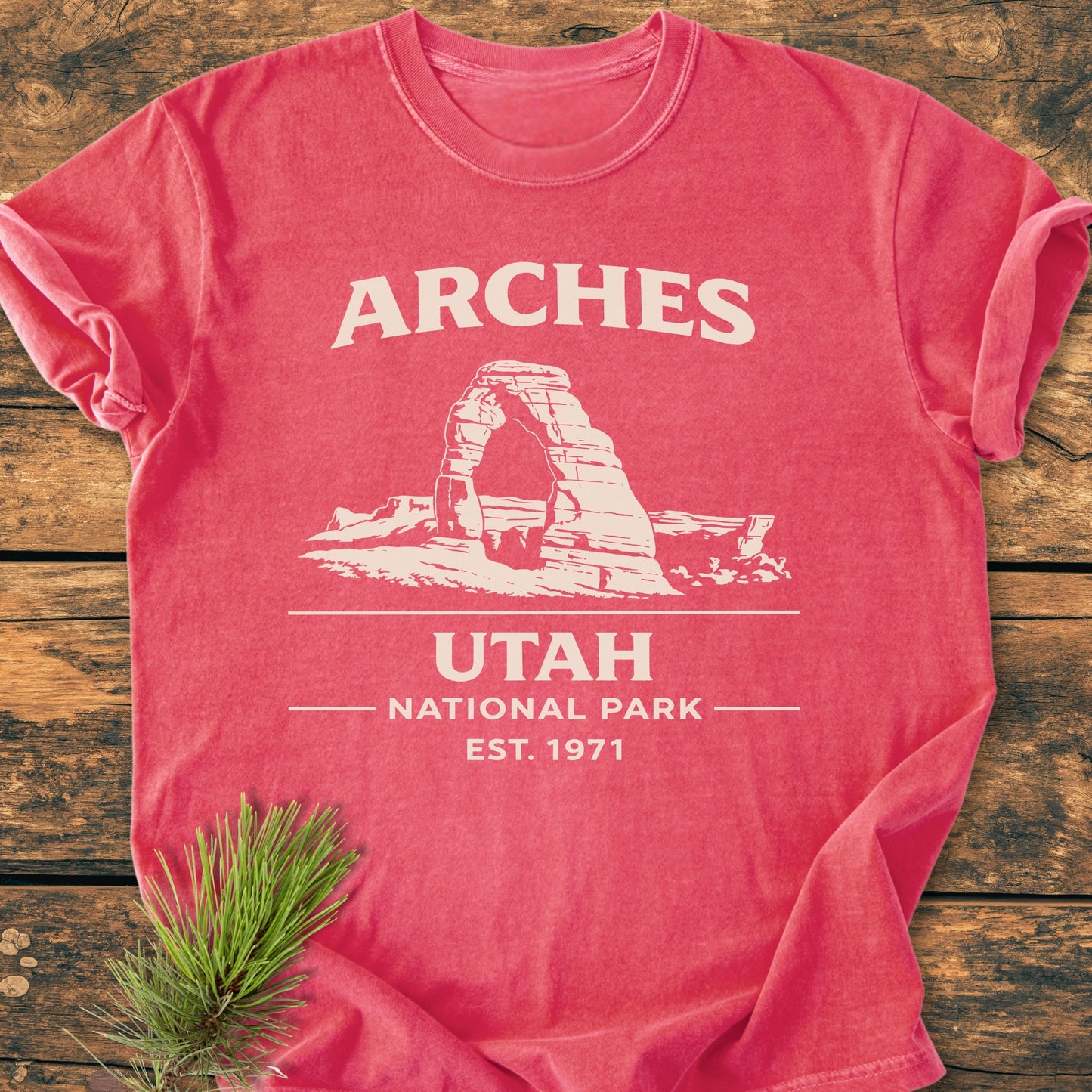 Arches National Park - Vintage Wash Tee featuring a white stone arch graphic and text. Displayed on a wooden surface with a pine sprig, highlighting its rustic, cozy appeal and relaxed fit.