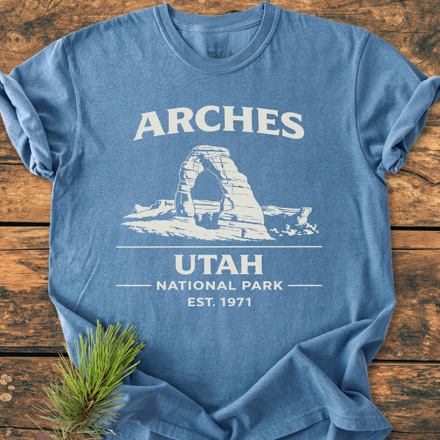 Arches National Park - Vintage Wash Tee in heather-blue featuring a white Delicate Arch sketch and text. Laid flat on wood, small evergreen sprig nearby. Relaxed fit, garment-dyed, ring-spun cotton.