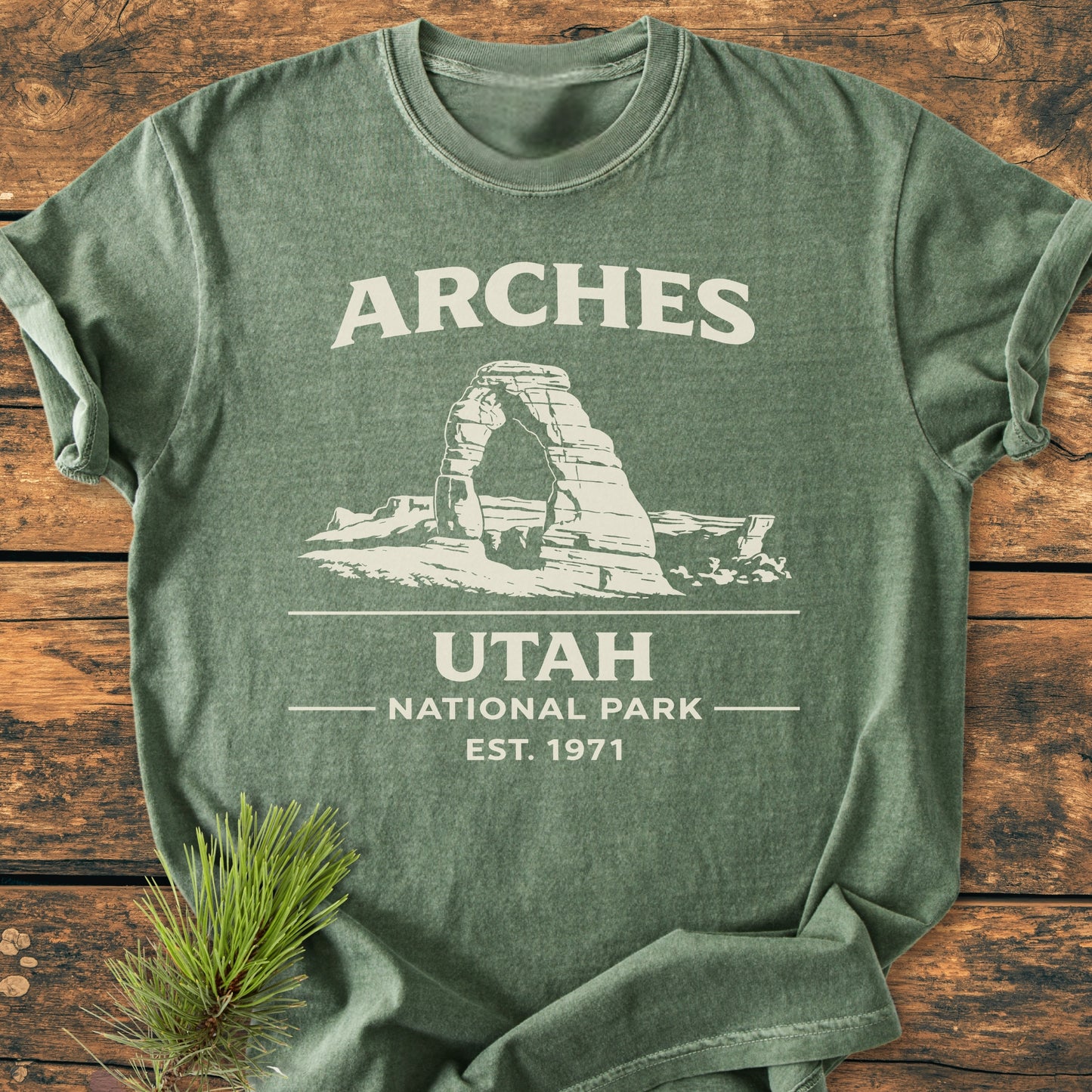 Arches National Park - Vintage Wash Tee laid flat, featuring ARCHES and a Delicate Arch drawing. Accompanied by a pine sprig, highlighting its relaxed fit and durable design.