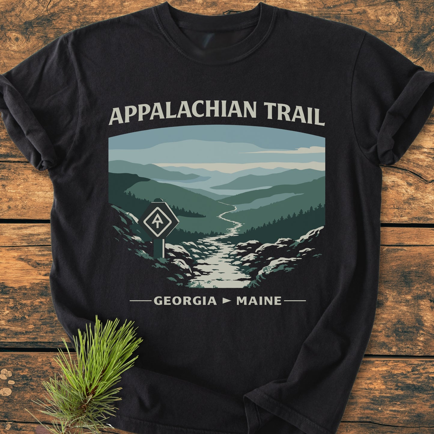 Appalachian Trail - Vintage Wash Tee featuring a stylized graphic of mountain ridges and trailblaze marker on a black T-shirt, laid on a wooden surface, with a pine sprig beside it.