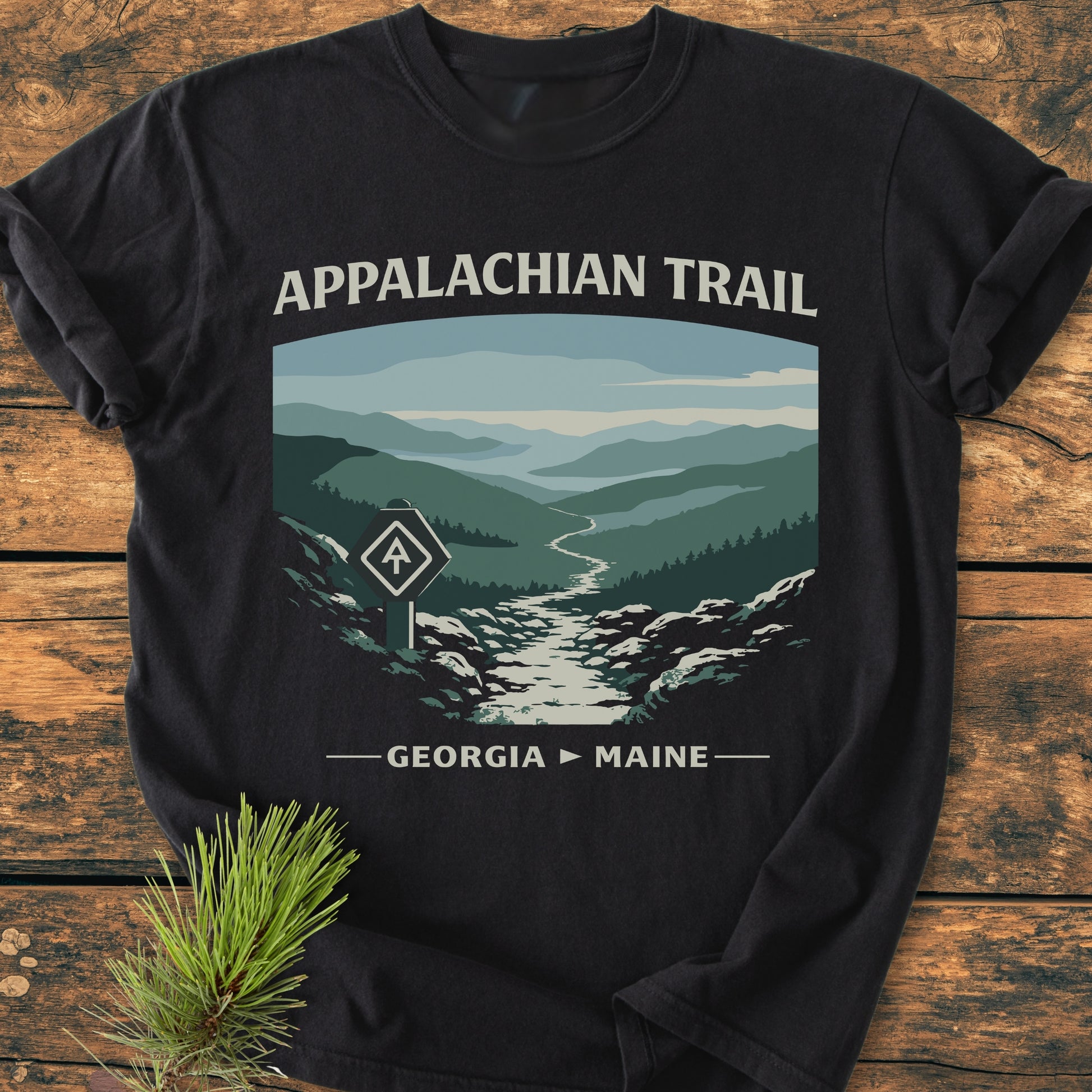 Appalachian Trail - Vintage Wash Tee featuring a stylized graphic of mountain ridges and trailblaze marker on a black T-shirt, laid on a wooden surface, with a pine sprig beside it.