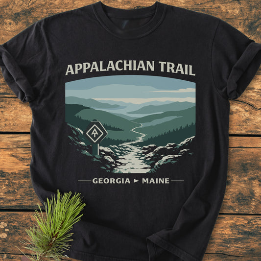 Appalachian Trail - Vintage Wash Tee featuring a stylized graphic of mountain ridges and trailblaze marker on a black T-shirt, laid on a wooden surface, with a pine sprig beside it.