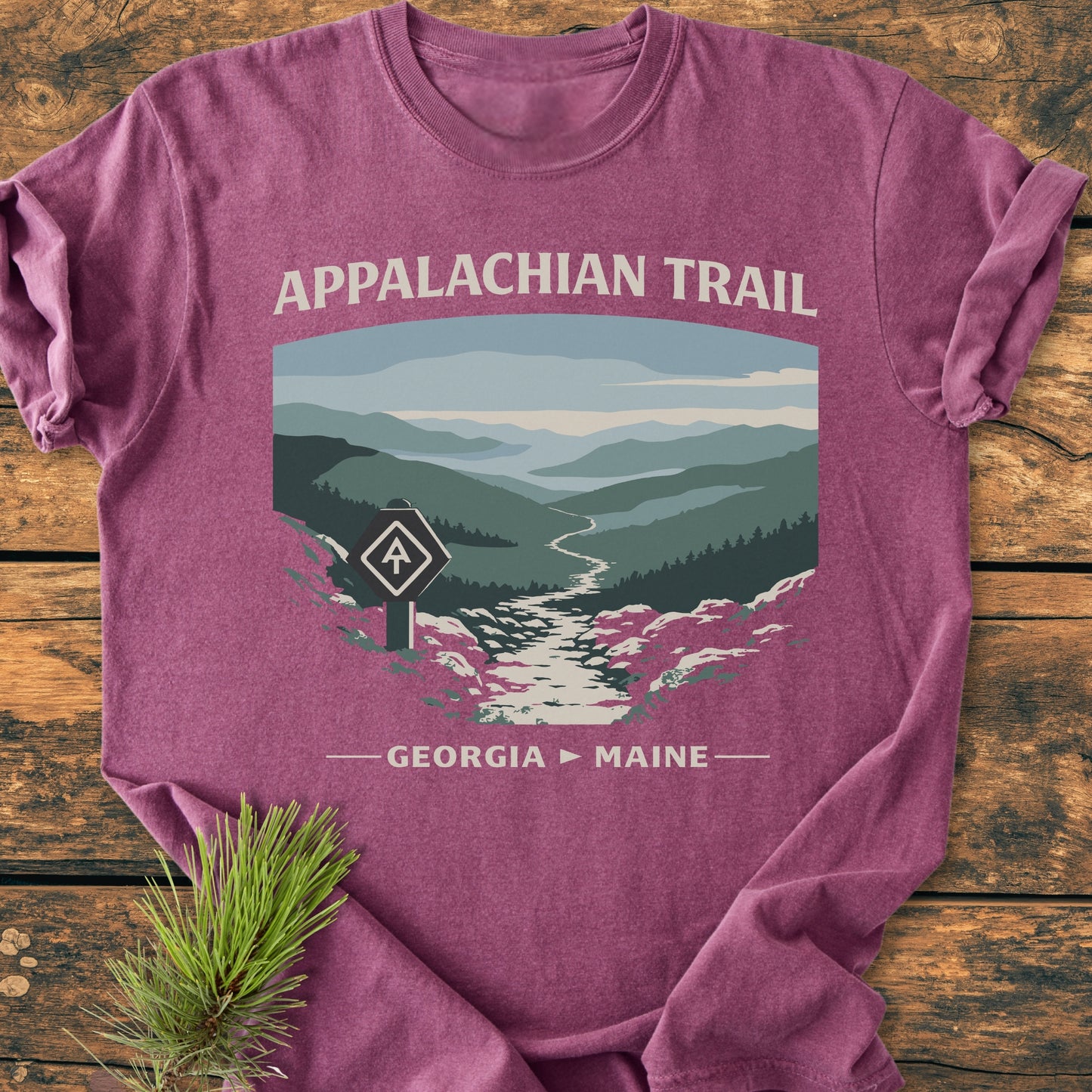 Appalachian Trail - Vintage Wash Tee features a mountain trail graphic with APPALACHIAN TRAIL and GEORGIA ► MAINE text, showcasing its rugged appeal on a wooden surface with a pine sprig beside.