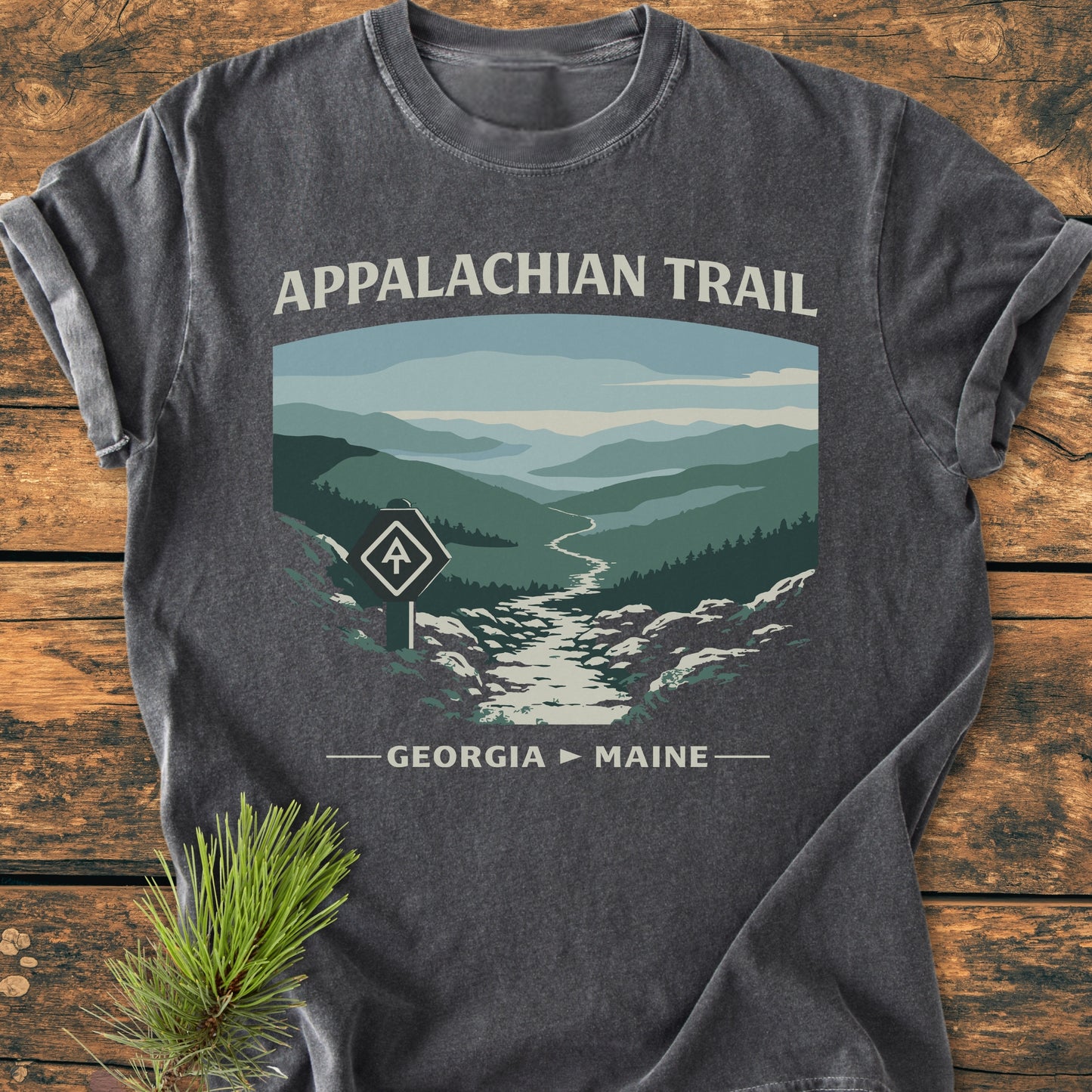 Appalachian Trail - Vintage Wash Tee features a stylized mountain landscape with trail and AT blaze, text reading “APPALACHIAN TRAIL” and “GEORGIA ► MAINE,” on dark gray fabric laid on wood.