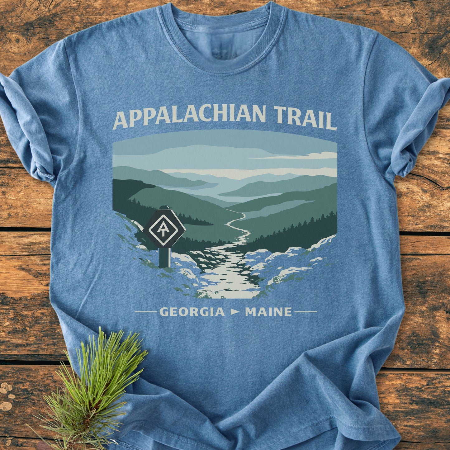 Appalachian Trail - Vintage Wash Tee: Heather-blue T-shirt featuring a mountain illustration and Appalachian Trail blaze, laid on a wooden surface with a pine sprig, emphasizing outdoor adventure.
