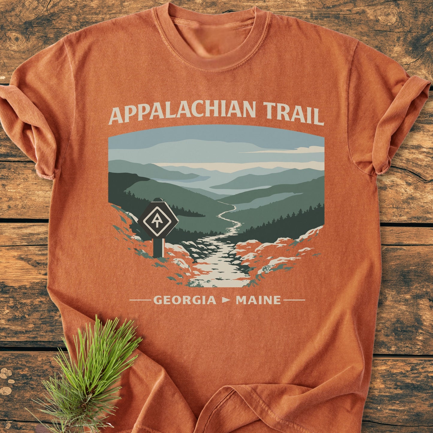 Appalachian Trail - Vintage Wash Tee features a stylized mountain landscape with a trail and signpost, on a rust-orange shirt, laid flat on wood, emphasizing its relaxed, durable design.