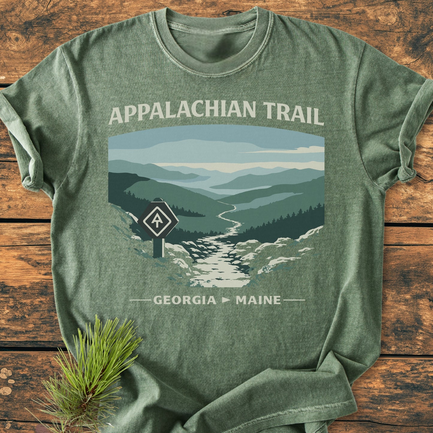 Appalachian Trail - Vintage Wash Tee: Forest-green T-shirt featuring an Appalachian Trail design with mountain scenery and trail-blaze sign, laid on a wooden surface with a pine sprig.