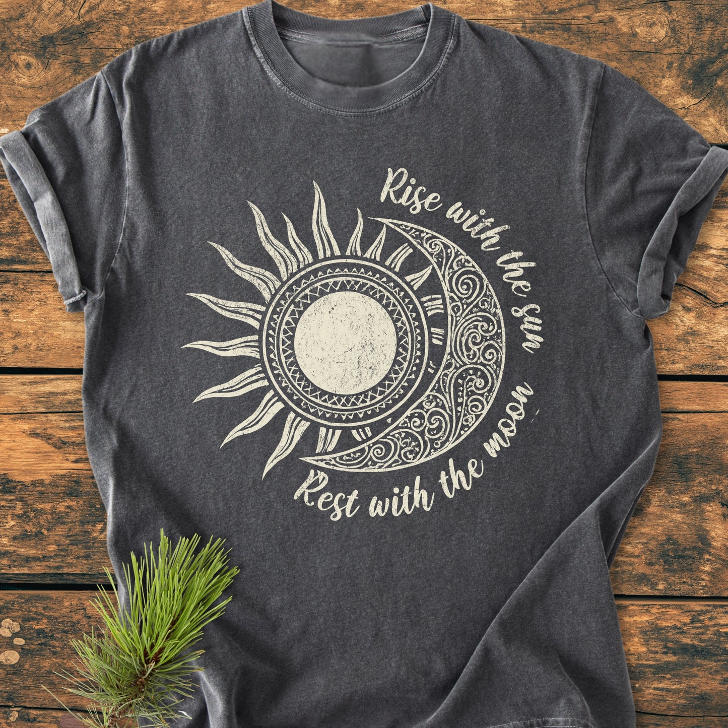 Sun & Moon Mystical Boho - Vintage Wash Tee featuring a stylized sun-and-moon graphic and Rise with the sun – Rest with the moon phrase, laid on a rustic wooden surface with a pine sprig accent.