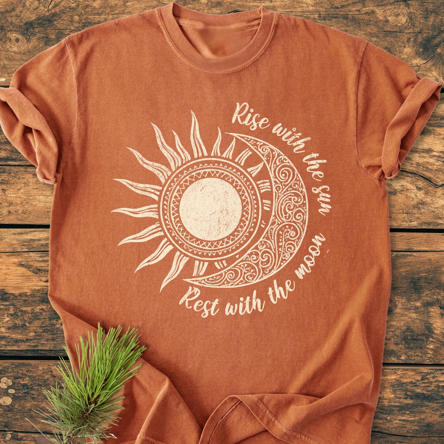 Sun & Moon Mystical Boho - Vintage Wash Tee with a sun and moon graphic, laid on wood, featuring Rise with the sun and Rest with the moon text, accented by a pine sprig.