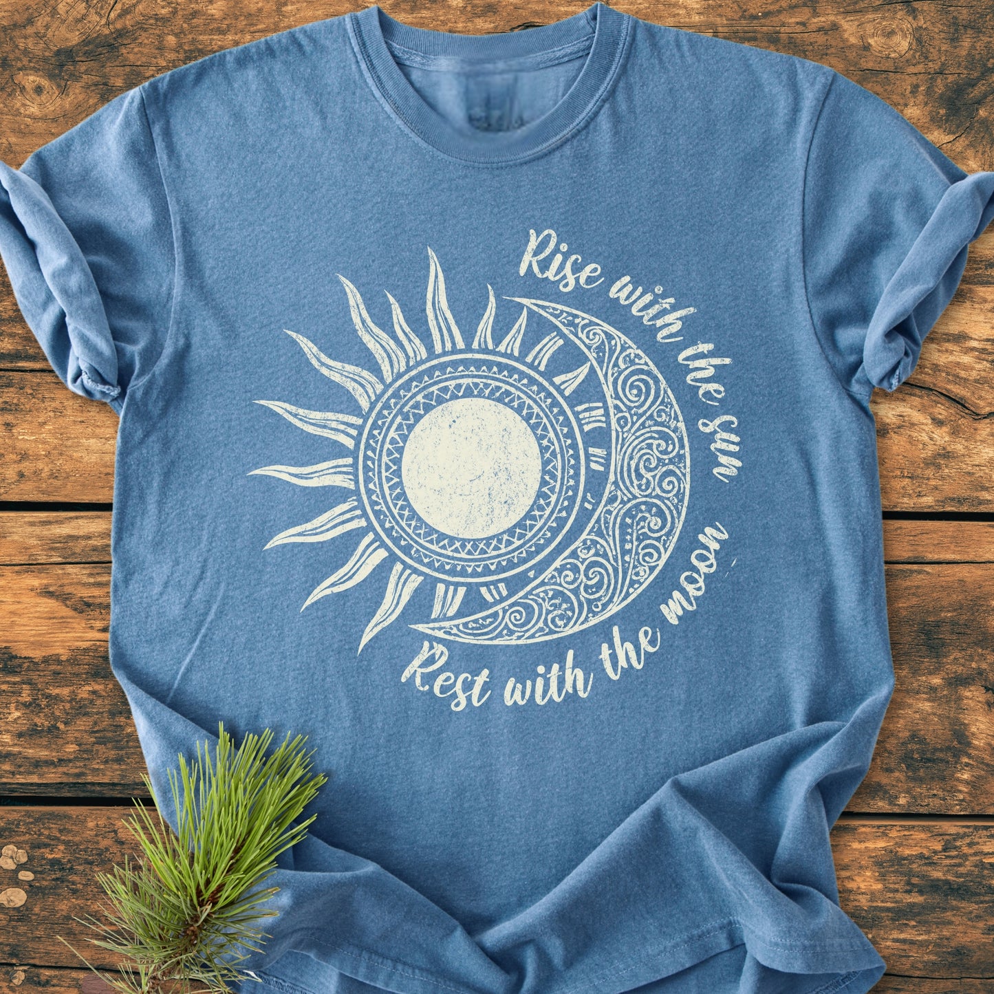Sun & Moon Mystical Boho - Vintage Wash Tee features a distressed white sun-moon graphic with Rise with the sun, Rest with the moon text, laid on a rustic wood surface.