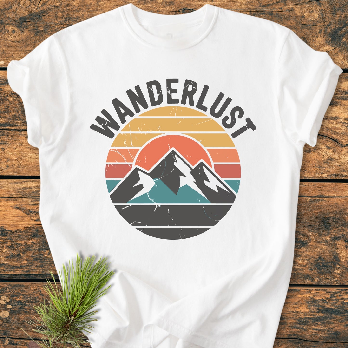 Wanderlust - Vintage Wash Tee featuring a retro mountain graphic on white cotton, laid on wood. Includes a pine sprig accent. Cozy, relaxed fit, ideal for everyday wear.