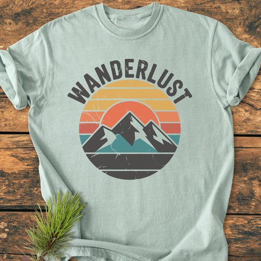 Wanderlust - Vintage Wash Tee: Light-colored T-shirt with retro-style sun and mountain graphic, featuring WANDERLUST text. Laid on wood surface, highlighting its relaxed fit and soft-washed, garment-dyed fabric.