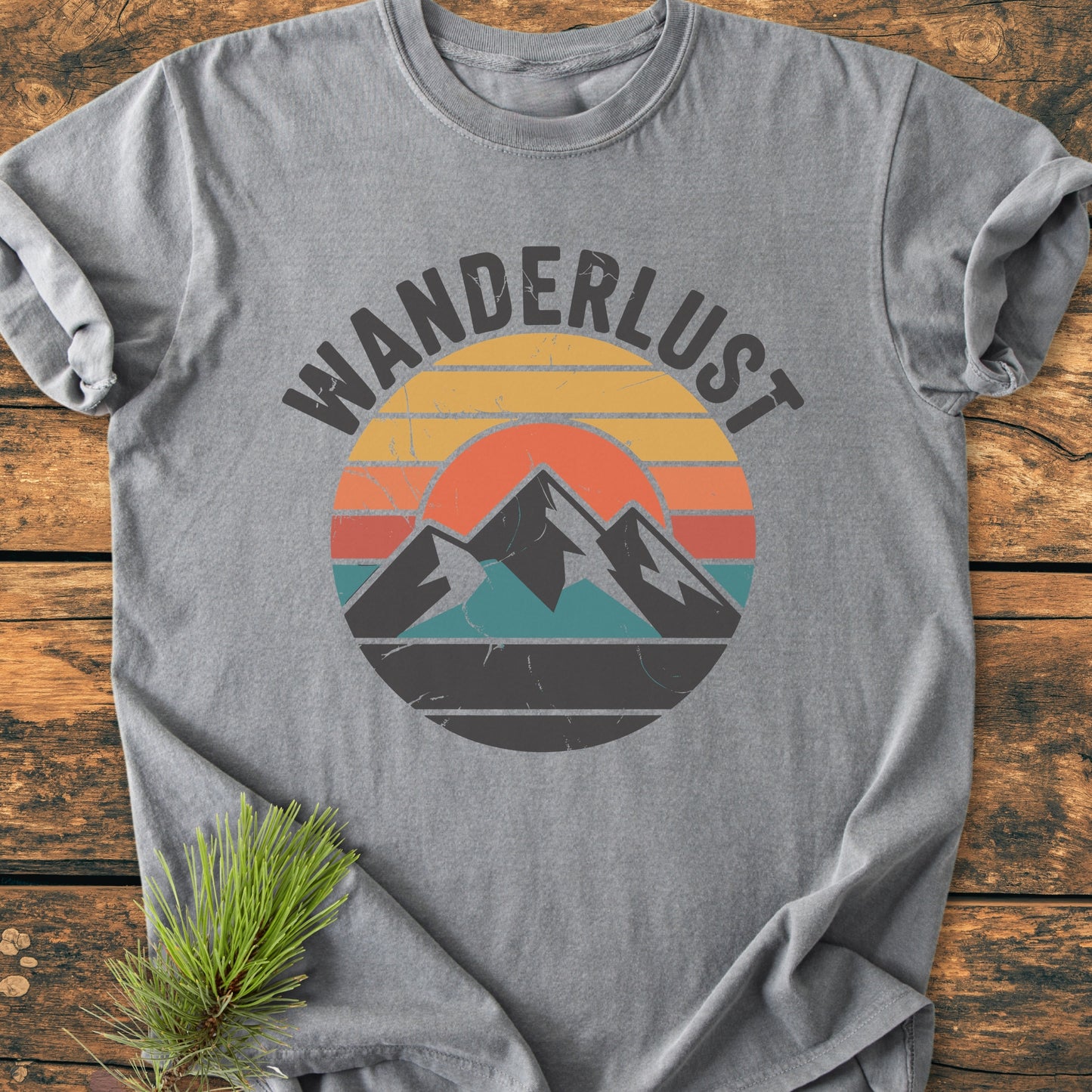 Wanderlust - Vintage Wash Tee: A heather-gray T-shirt featuring a retro mountain graphic and WANDERLUST text, laid on a wooden surface with a pine branch accentuating the outdoor theme.