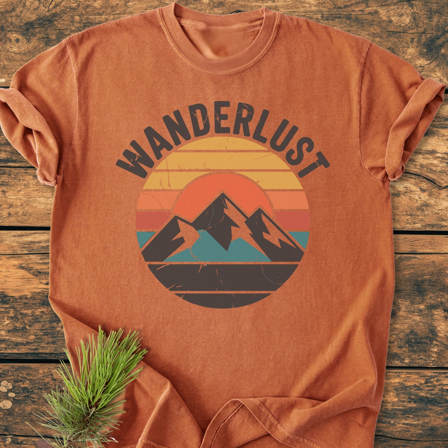 Wanderlust - Vintage Wash Tee: A rust-orange, vintage-washed T-shirt with a retro sunset and mountain graphic, laid on wood, pine sprig accents. Perfect for comfort and style with durable, garment-dyed fabric.