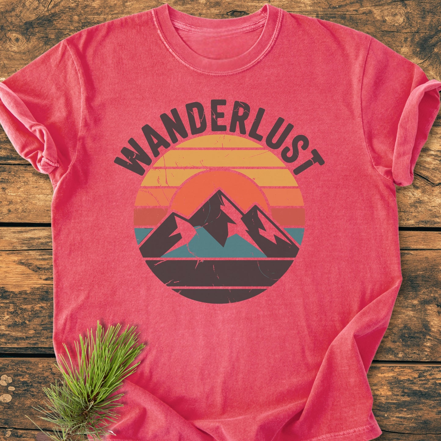 Wanderlust - Vintage Wash Tee featuring a WANDERLUST graphic with a sun and mountains, laid on wood. Sprig of pine needles adds a rustic touch.