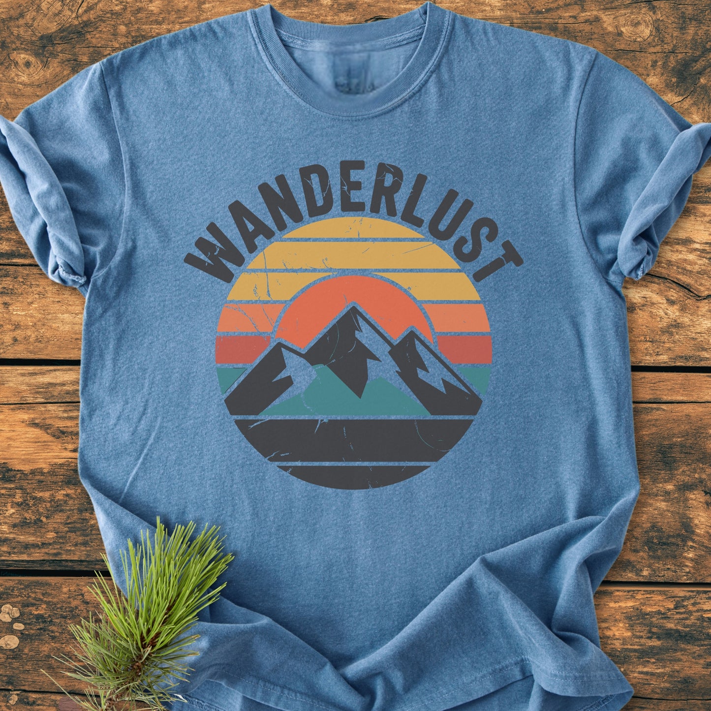 Wanderlust - Vintage Wash Tee: Heathered mid-blue T-shirt with retro mountain graphic and WANDERLUST text, laid on wood. Cozy, relaxed fit, durable garment-dyed fabric with double-needle stitching.