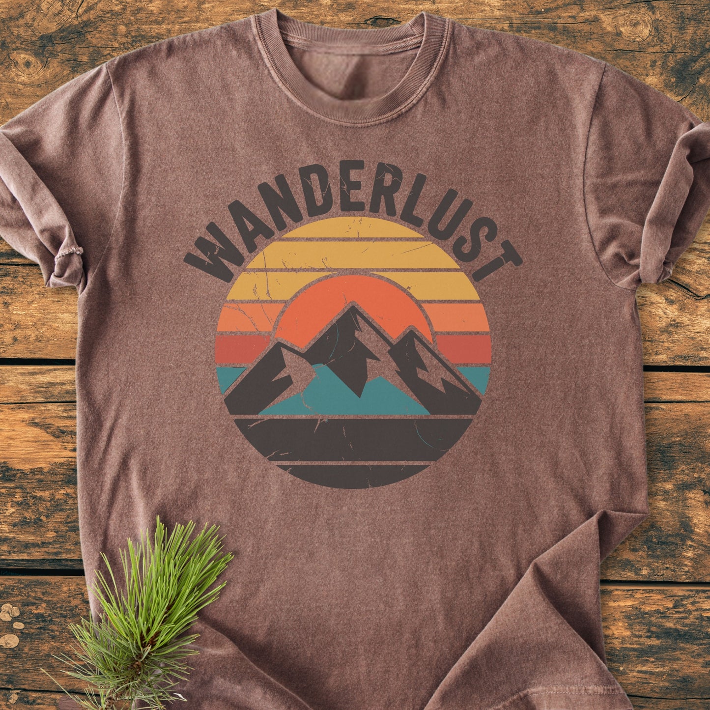 Wanderlust - Vintage Wash Tee, a brown short-sleeve shirt with a circular graphic of sunset stripes and mountains, displayed flat on wood, showcasing its relaxed fit and durable design.