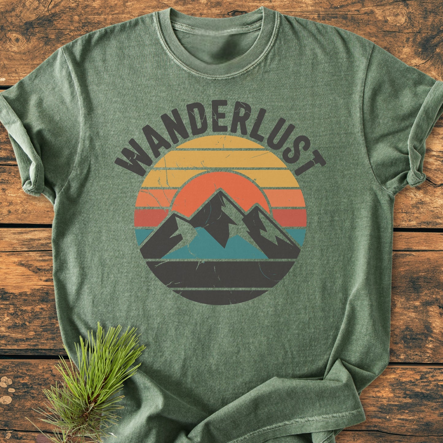 Wanderlust - Vintage Wash Tee with a vintage sunset and mountain graphic on soft green fabric, laid on a wooden surface, showcasing its relaxed fit and durable, ring-spun cotton construction.