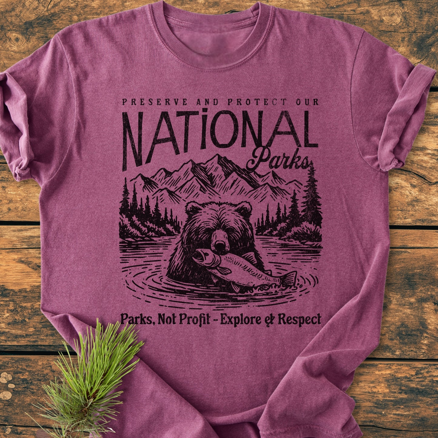 Preserve Our National Parks - Vintage Wash Tee: Heathered berry-purple T-shirt featuring a bold nature-conservation design with a bear illustration, emphasizing environmental protection. Laid flat on a wooden surface with pine sprig accent.