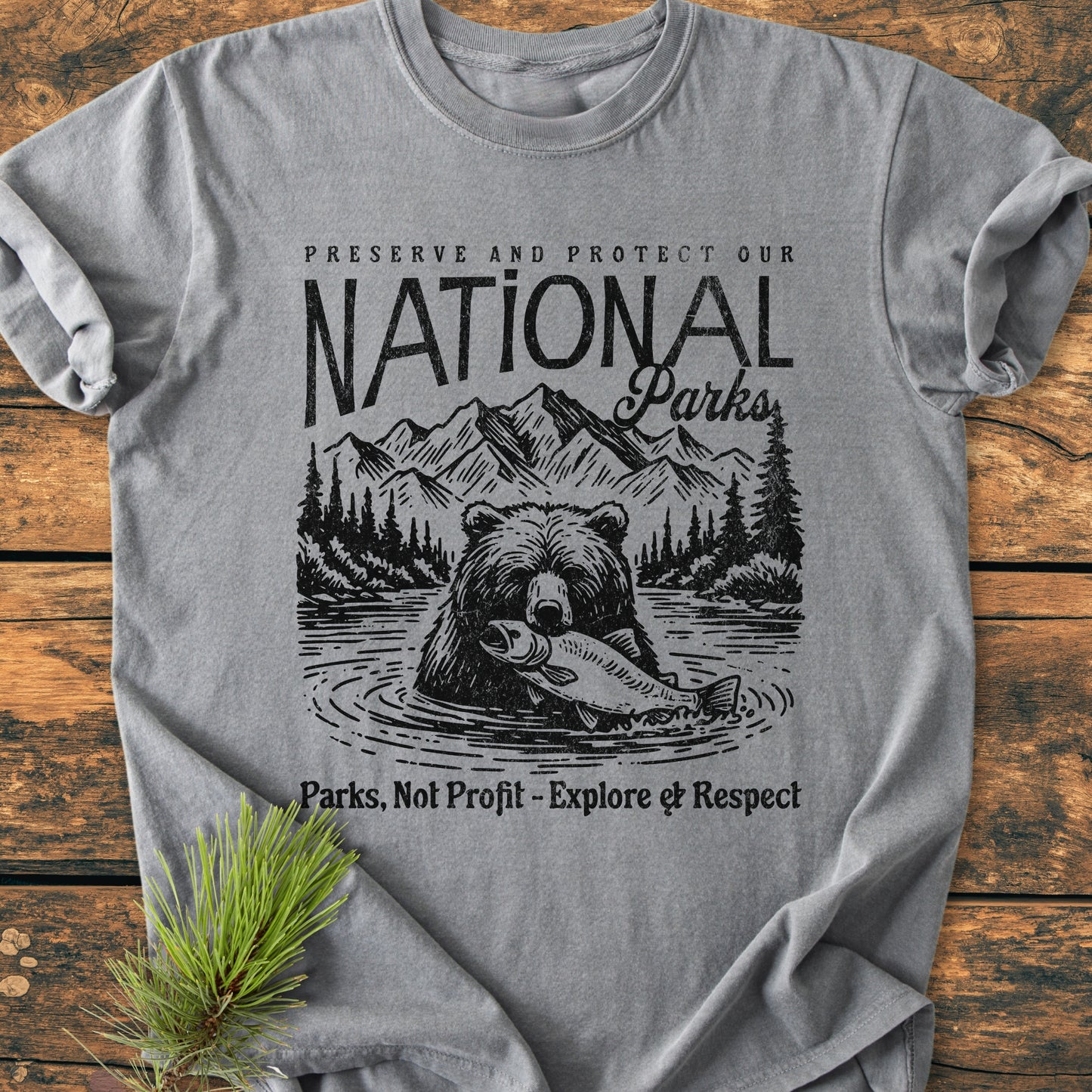 Preserve Our National Parks - Vintage Wash Tee features a bear with a fish, mountain range, and pine trees, emphasizing Parks, Not Profit. Heather gray, laid on wood, with a pine branch accent.