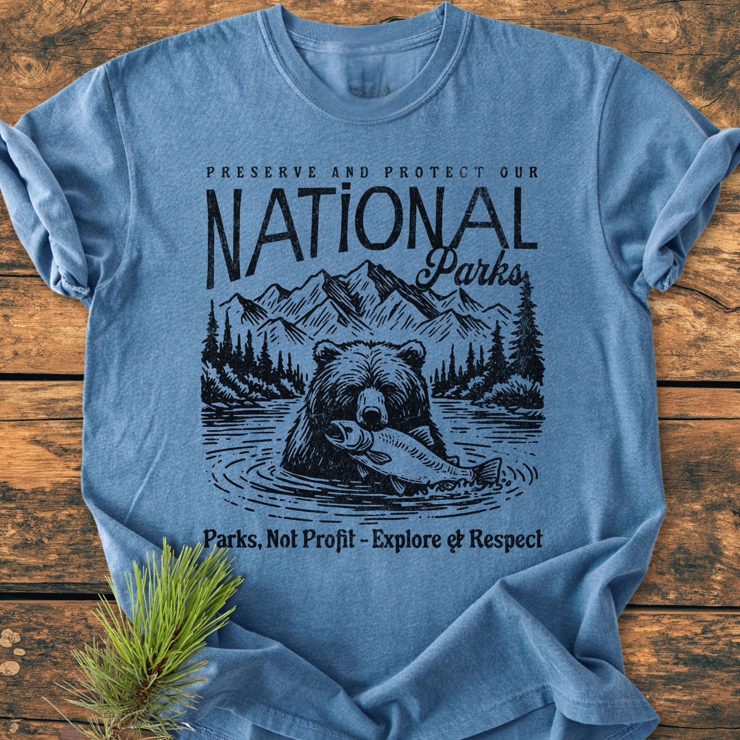Preserve Our National Parks - Vintage Wash Tee features a bear and nature scene with conservation messages, laid on a wood surface, highlighting its relaxed fit and durable, soft-washed 100% cotton fabric.