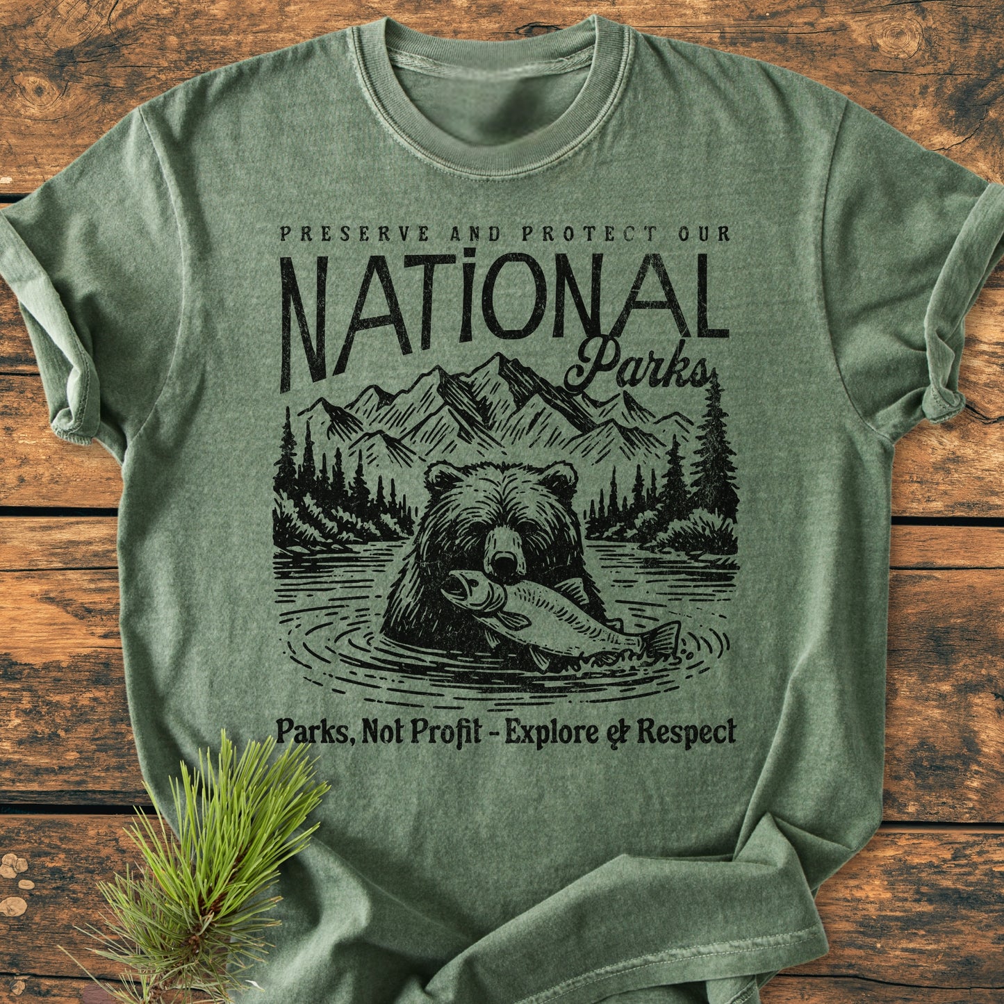 Preserve Our National Parks - Vintage Wash Tee, featuring a black, vintage-style graphic of a bear with fish, pine trees, and mountains, laid flat on wood with a pine sprig beside it.