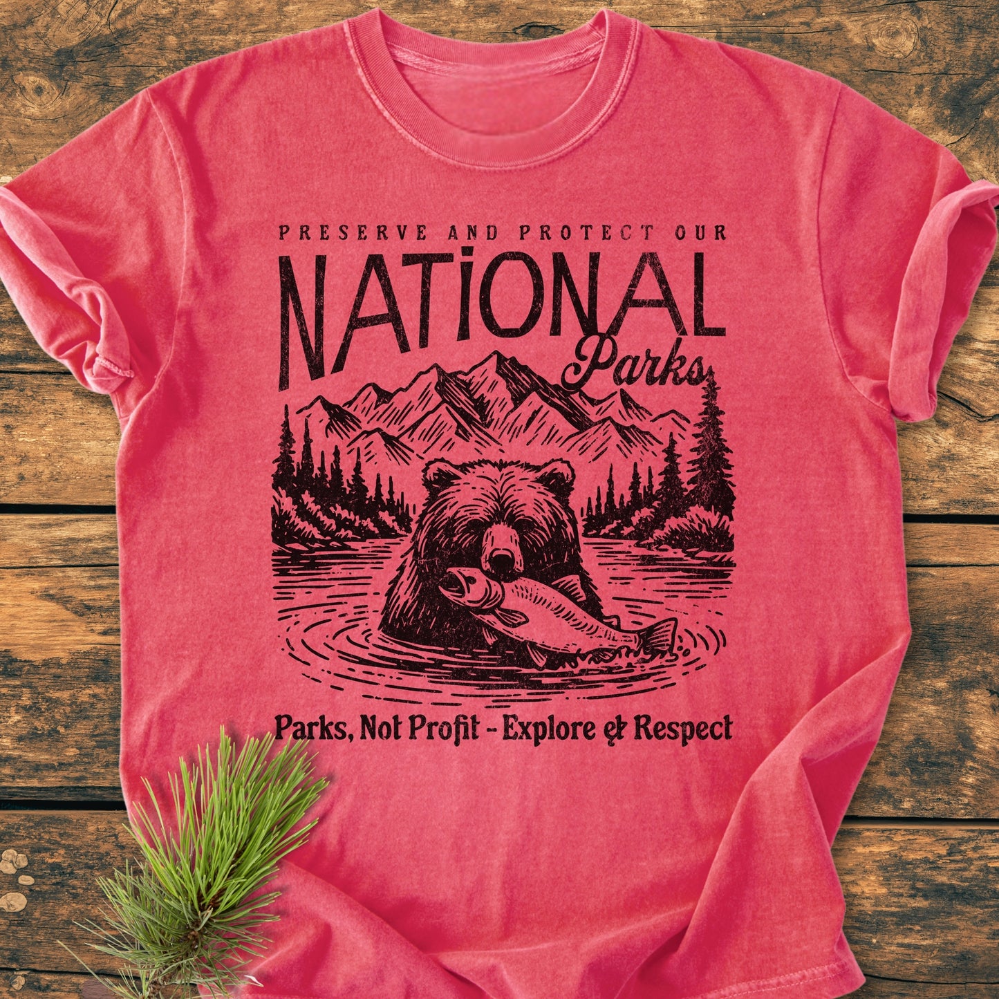 Preserve Our National Parks - Vintage Wash Tee features a bear in a river design with mountains and trees, promoting park conservation. Laid on wood, it includes a pine sprig accent.