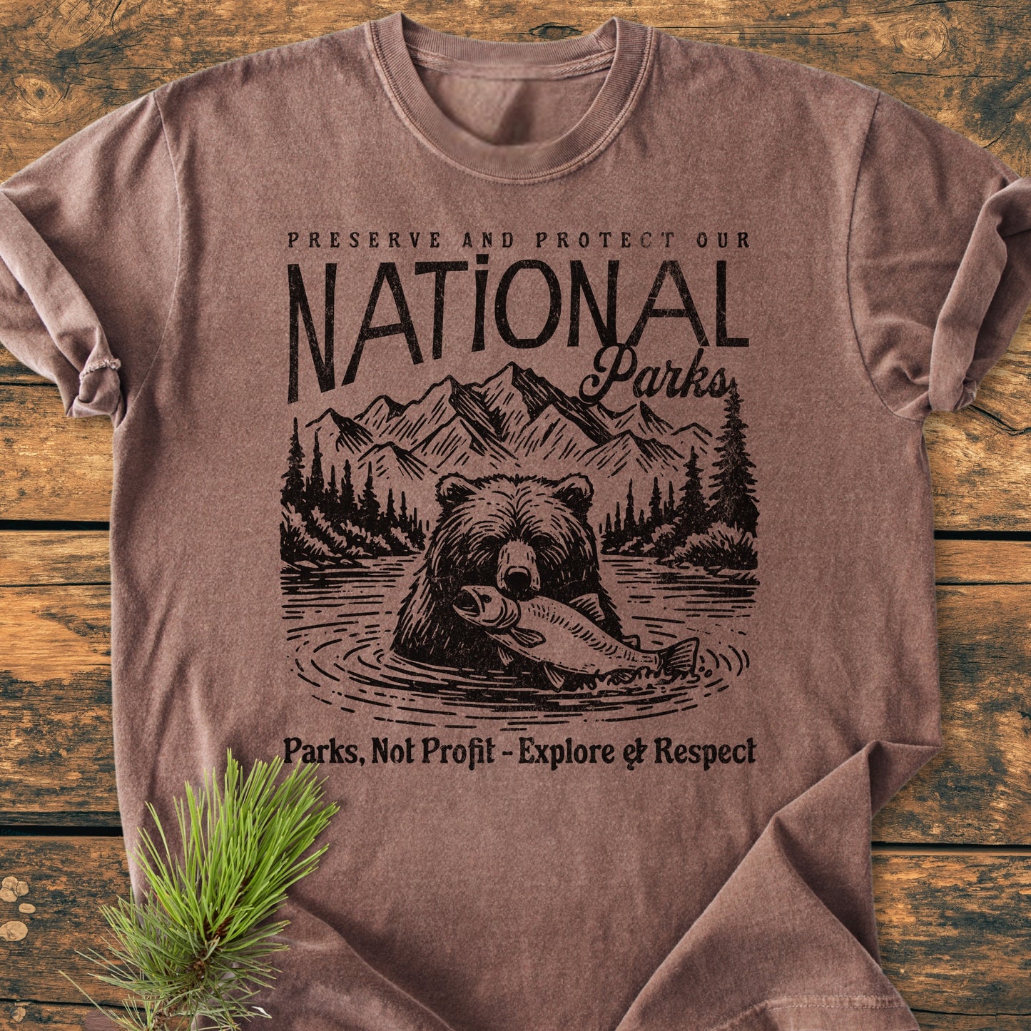 Preserve Our National Parks - Vintage Wash Tee featuring a bear and nature design, laid on wood. Highlights Preserve and Protect Our National Parks and Parks, Not Profit – Explore & Respect. Pine branch beside it.