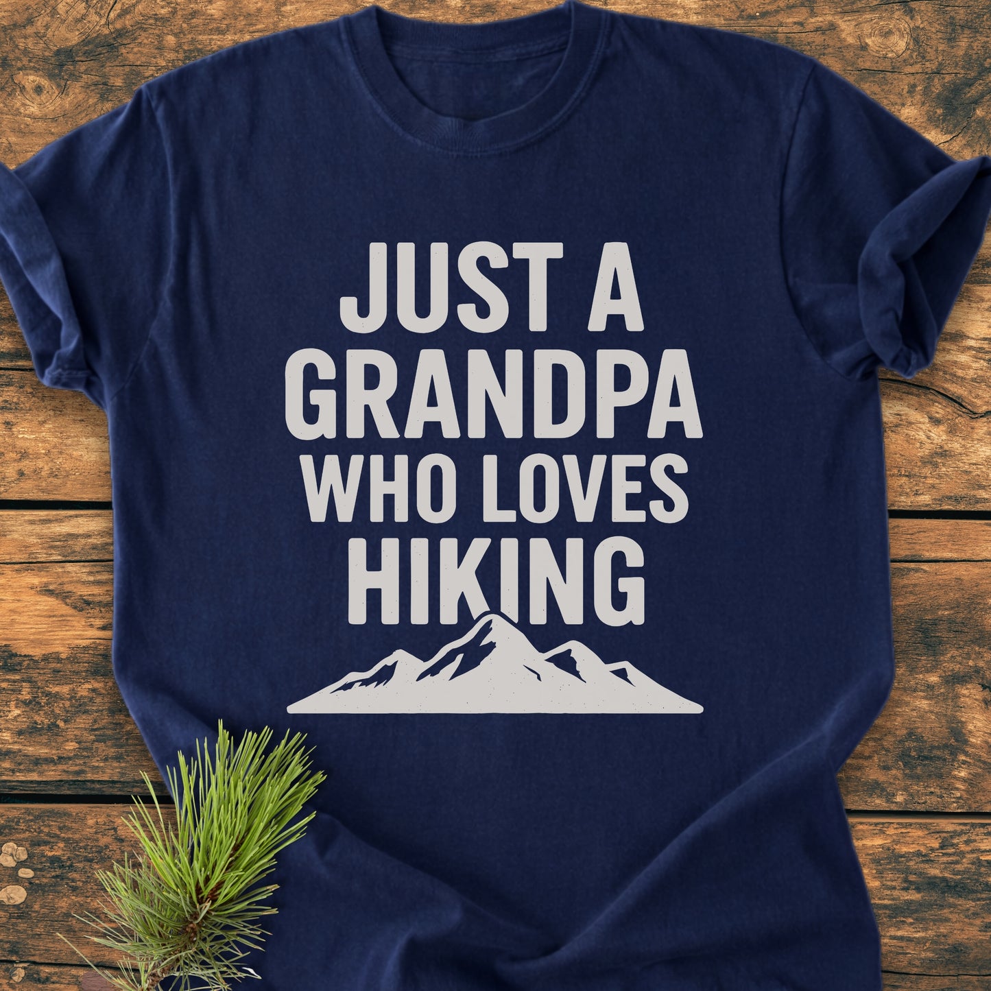 Hiking Grandpa - Vintage Wash Tee laid flat showing JUST A GRANDPA WHO LOVES HIKING in white with a mountain silhouette, styled with a pine sprig on wooden surface.