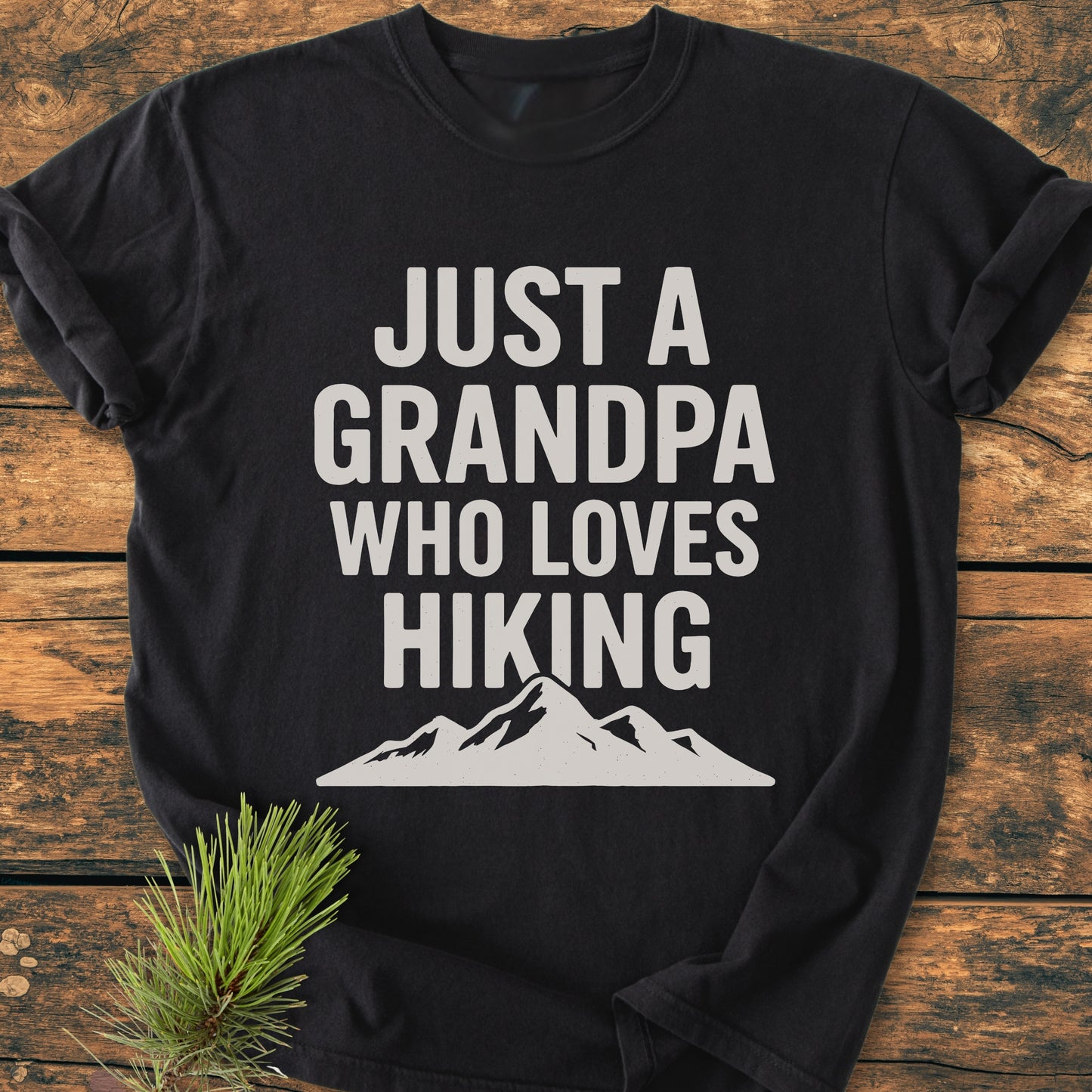 Hiking Grandpa - Vintage Wash Tee: Black T-shirt with JUST A GRANDPA WHO LOVES HIKING in white, featuring a mountain graphic. Displayed on wood with pine needles, highlighting its relaxed fit and cozy material.