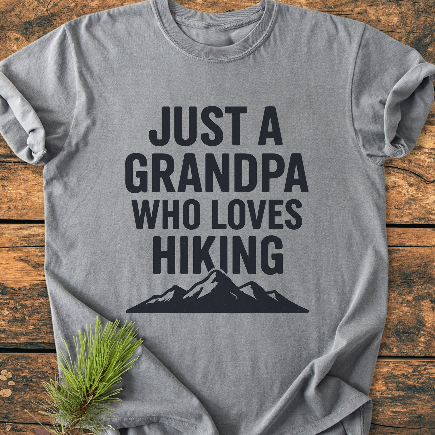 Hiking Grandpa - Vintage Wash Tee: Heather-gray T-shirt with JUST A GRANDPA WHO LOVES HIKING text and mountain silhouette, laid flat with pine branch accentuating its outdoorsy theme.