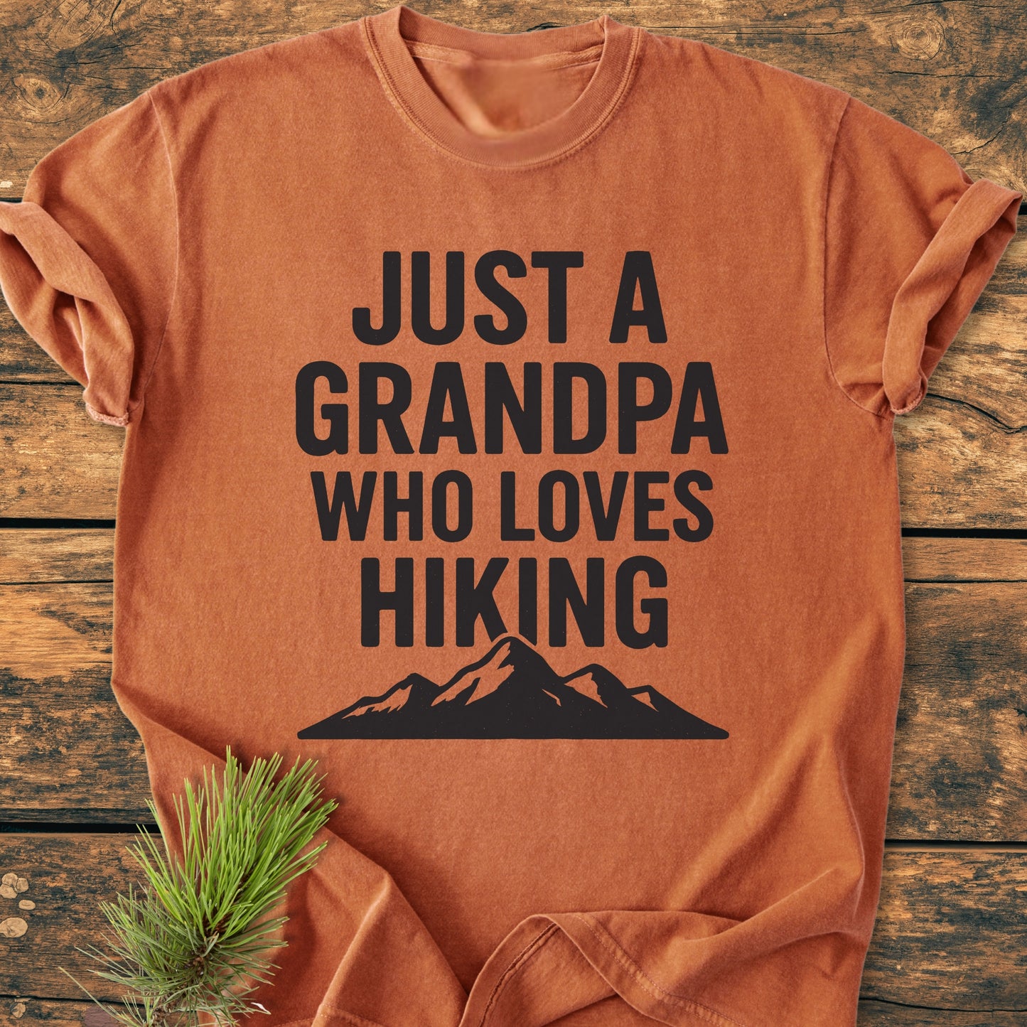 Hiking Grandpa - Vintage Wash Tee: Burnt-orange T-shirt with JUST A GRANDPA WHO LOVES HIKING in bold black, mountain silhouette below, sleeves rolled, displayed on wood, pine sprig accent.