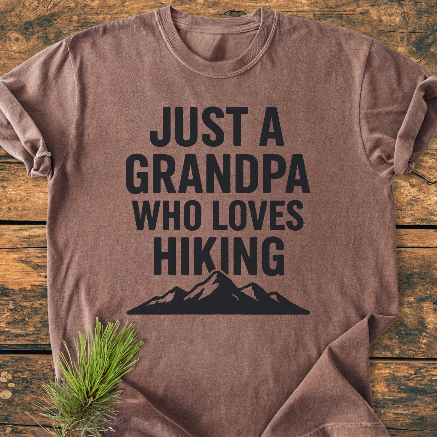 Hiking Grandpa - Vintage Wash Tee displayed on wood, featuring JUST A GRANDPA WHO LOVES HIKING in black with a mountain silhouette. Pine needles adorn the corner, highlighting its rustic charm.