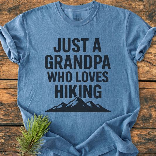Hiking Grandpa - Vintage Wash Tee on wooden surface, featuring bold slogan “JUST A GRANDPA WHO LOVES HIKING” with mountain silhouette. Pine sprig adds rustic touch. Comfortable, durable, and relaxed fit.
