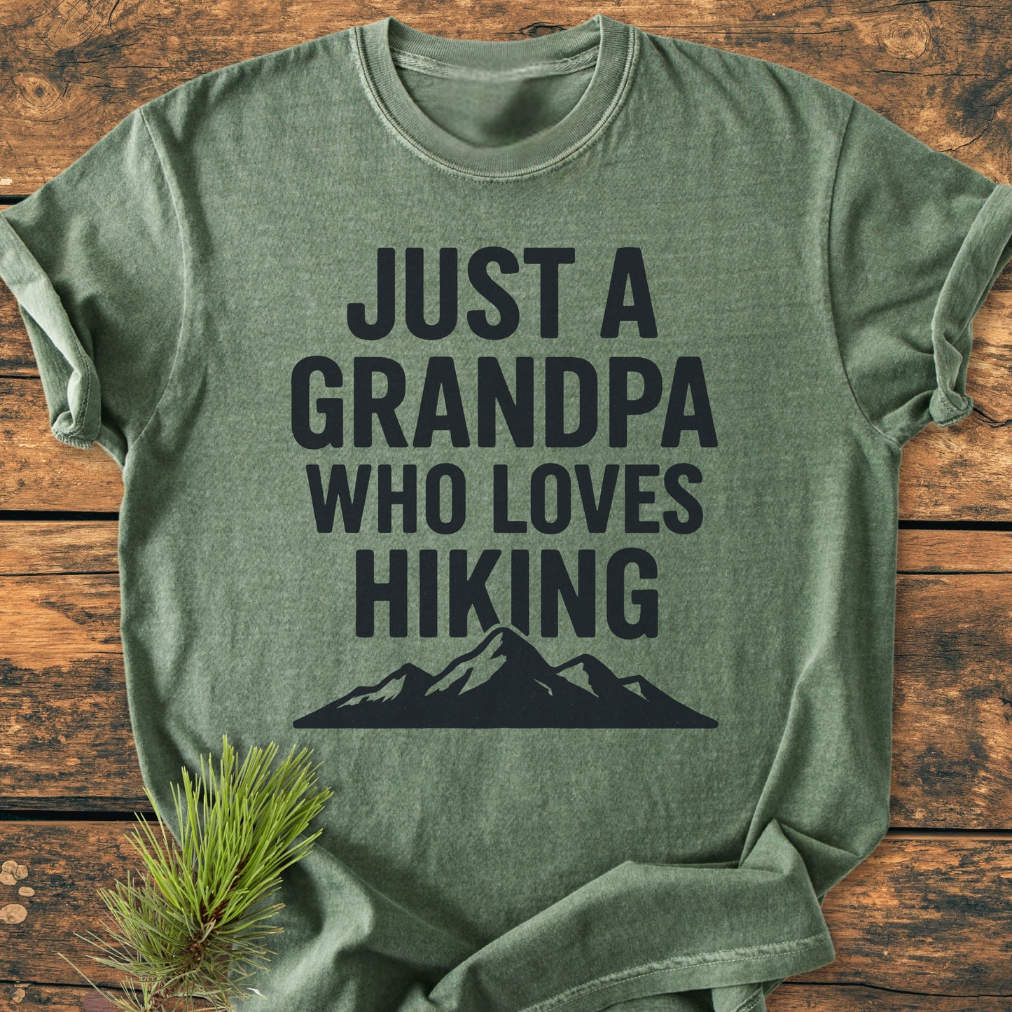 Heather-green Hiking Grandpa - Vintage Wash Tee with bold text and mountain silhouette, laid on rustic wood, accented by pine needles, showcasing an outdoorsy theme.