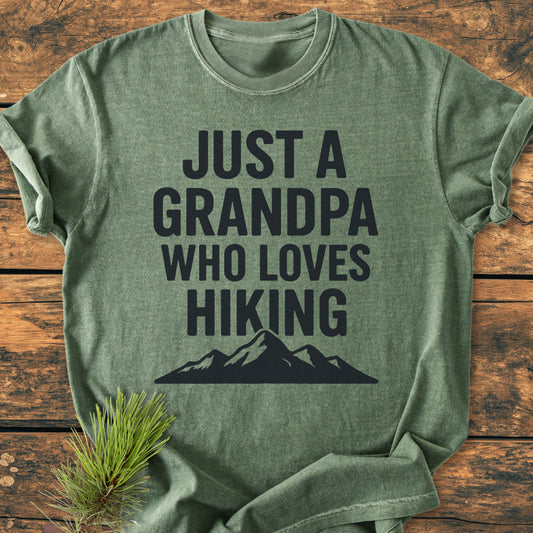 Heather-green Hiking Grandpa - Vintage Wash Tee with bold text and mountain silhouette, laid on rustic wood, accented by pine needles, showcasing an outdoorsy theme.