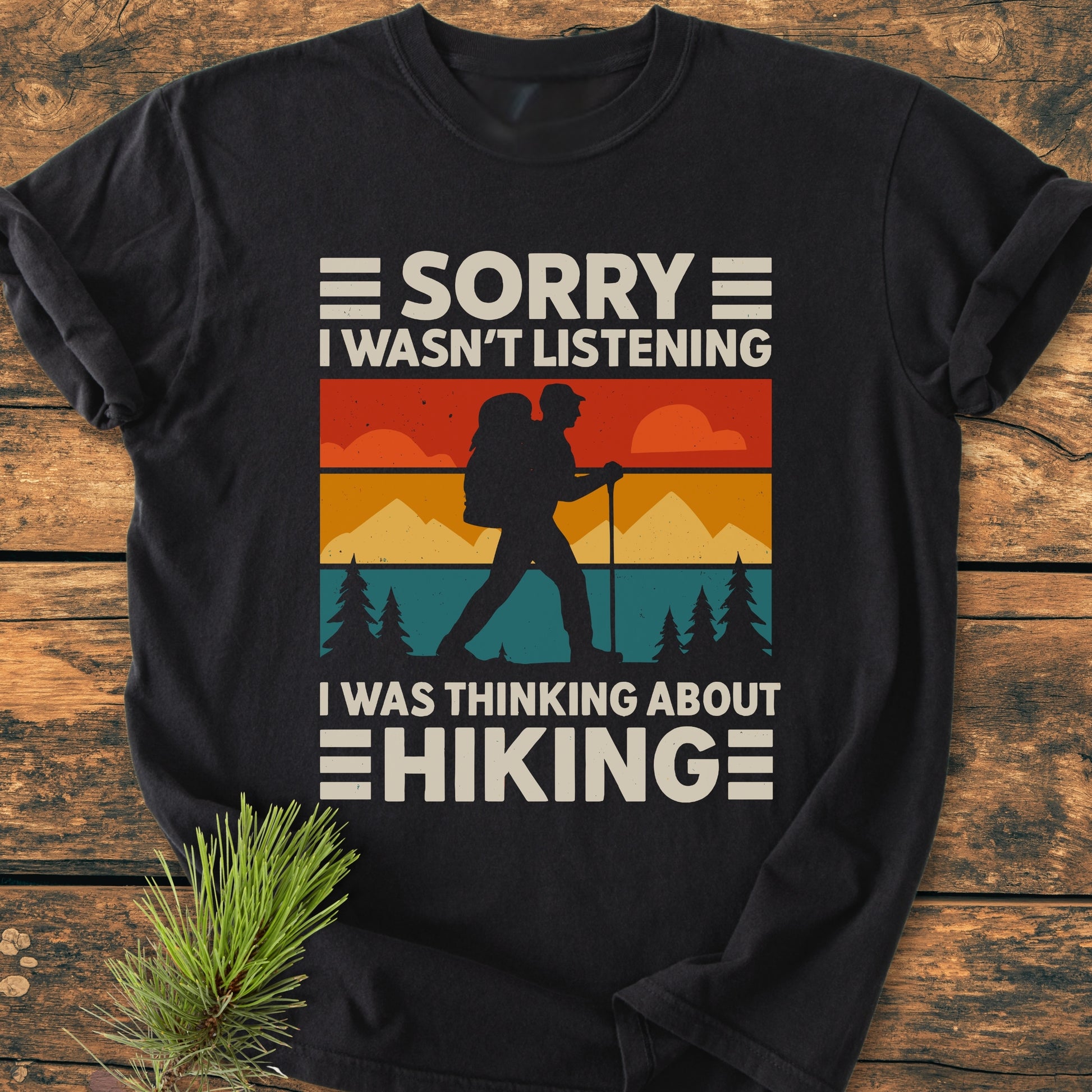 Thinking About Hiking - Vintage Wash Tee featuring a retro hiker graphic with vibrant color bands and hiking slogan, laid on wood with a pine sprig, highlighting its relaxed fit and durability.