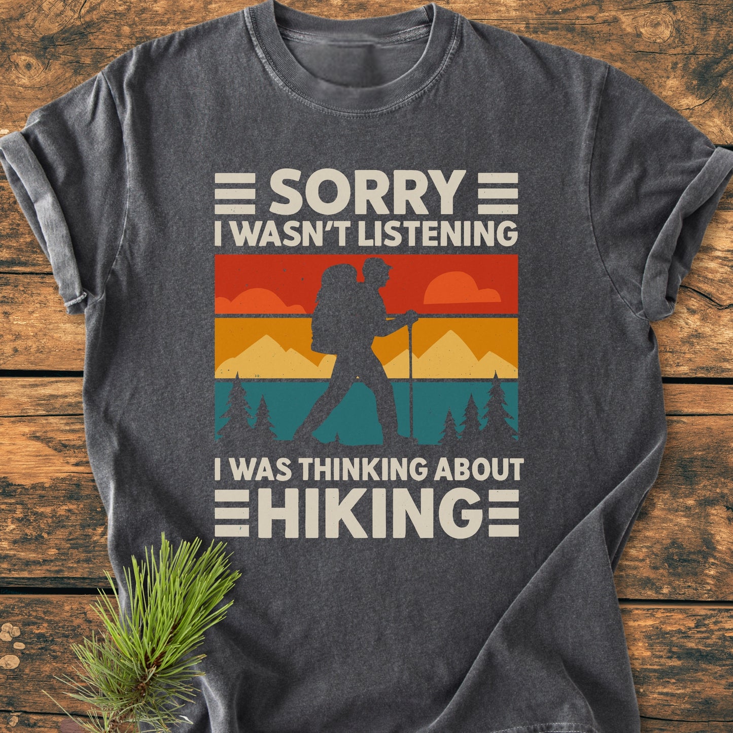 Thinking About Hiking - Vintage Wash Tee features a retro landscape graphic with a backpacker, printed on a charcoal-gray shirt, laid on wood with a pine sprig for a cozy, outdoorsy vibe.