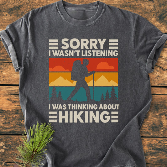 Thinking About Hiking - Vintage Wash Tee features a retro landscape graphic with a backpacker, printed on a charcoal-gray shirt, laid on wood with a pine sprig for a cozy, outdoorsy vibe.